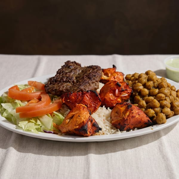 A white plate with grilled chicken, beef, rice, chickpeas, salad, and a side of green sauce.