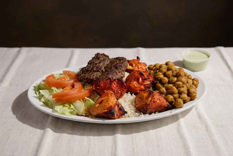 A white plate with grilled chicken, beef, rice, chickpeas, salad, and a side of green sauce.