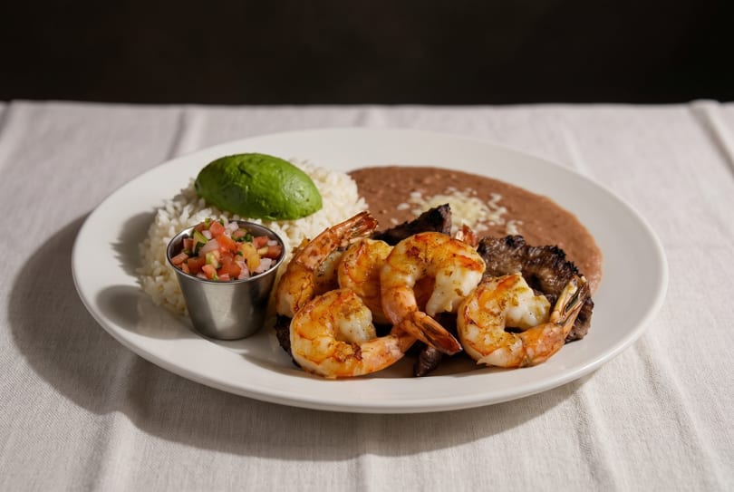 Grilled shrimp and steak with rice, refried beans, avocado, and pico de gallo on a white plate.