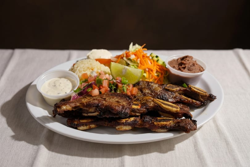 Grilled short ribs with rice, pico de gallo, salad, refried beans, and sauces on a white plate.