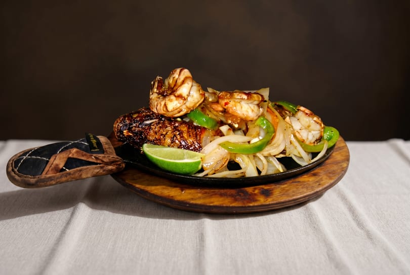 Sizzling shrimp and fish fajitas with onions, green peppers, and lime on a wooden platter.